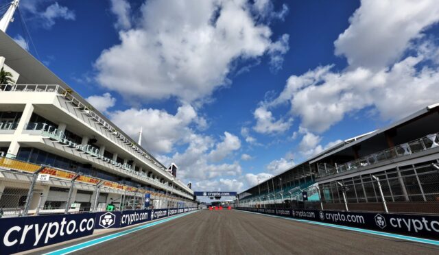 Wide view of a race track flanked by white pit buildings, sponsor banners reading 'crypto.com' along the barriers, under a bright blue sky with white clouds.