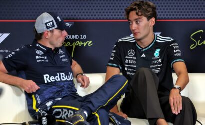 Two racing drivers sit on a white couch and converse at a press event; left in a Red Bull polo and cap, right in a black Mercedes shirt, backdrop shows sponsor logos and 'Singapore' text.