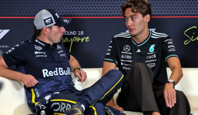 Two racing drivers sit on a white couch and converse at a press event; left in a Red Bull polo and cap, right in a black Mercedes shirt, backdrop shows sponsor logos and 'Singapore' text.