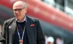 Smiling older man with glasses in a dark jacket and lanyard standing at a motorsport event, with a blurred red structure in the background.