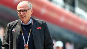 Smiling older man with glasses in a dark jacket and lanyard standing at a motorsport event, with a blurred red structure in the background.