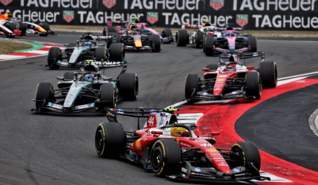 Motorsport race: multiple Formula 1 cars cornering on a track with red and white curbs, TAG Heuer banners in the background