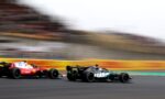 Three Formula 1 cars race at high speed on a track, with a red car on the left and a black Mercedes on the right, the stands blurred in the background.