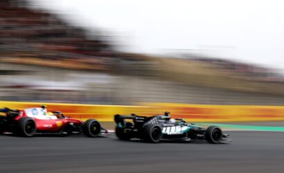 Three Formula 1 cars race at high speed on a track, with a red car on the left and a black Mercedes on the right, the stands blurred in the background.