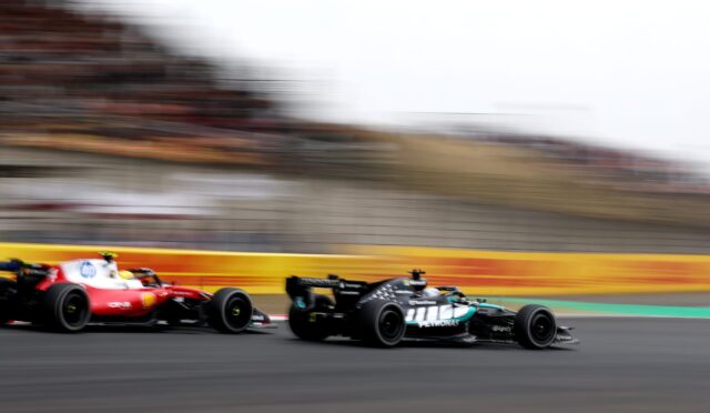 Three Formula 1 cars race at high speed on a track, with a red car on the left and a black Mercedes on the right, the stands blurred in the background.