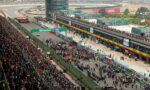 Aerial view of a Formula One race circuit with packed grandstands, a busy pit lane, and team tents along the pit wall.