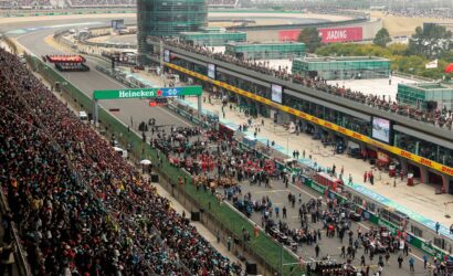 Aerial view of a Formula One race circuit with packed grandstands, a busy pit lane, and team tents along the pit wall.