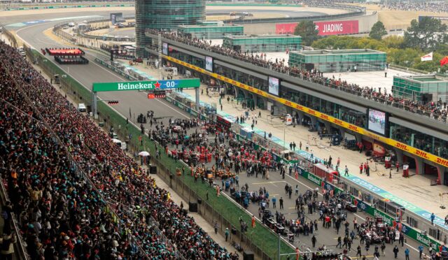 Aerial view of a Formula One race circuit with packed grandstands, a busy pit lane, and team tents along the pit wall.