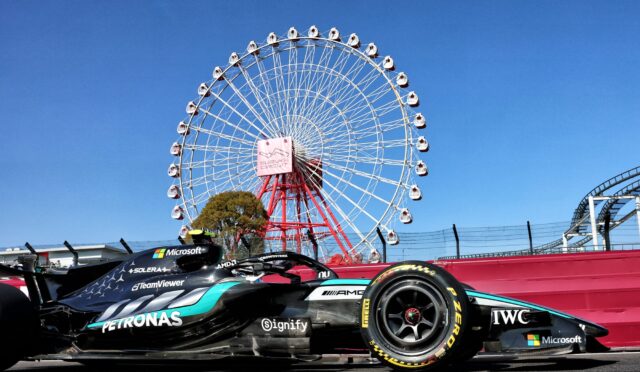 Formula 1 race car with Petronas livery on a racetrack, ferris wheel visible in the blue sky behind it.