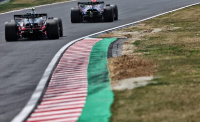 Rear view of two Formula 1 cars racing around a left-hand bend, with a red-and-white striped curb and green runoff on the inside of the track.