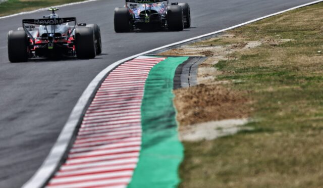Rear view of two Formula 1 cars racing around a left-hand bend, with a red-and-white striped curb and green runoff on the inside of the track.
