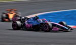 Pink-and-blue Formula 1 car cornering on a racetrack with another car blurred in the background