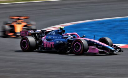 Pink-and-blue Formula 1 car cornering on a racetrack with another car blurred in the background