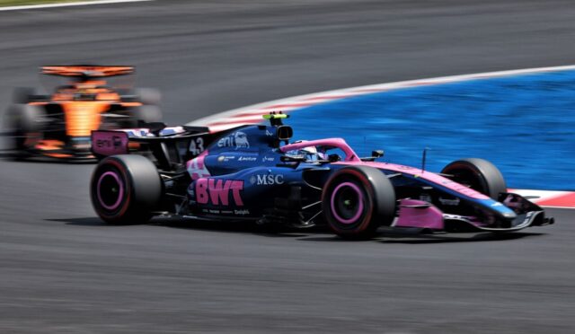 Pink-and-blue Formula 1 car cornering on a racetrack with another car blurred in the background