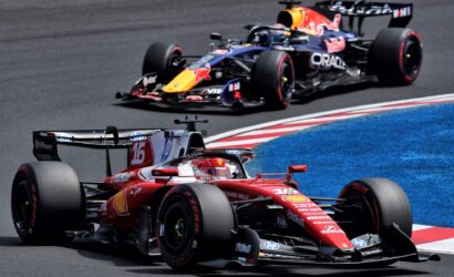 Red Ferrari Formula 1 car leading a race, closely followed by a Red Bull car on a curving track with blue run-off area behind.