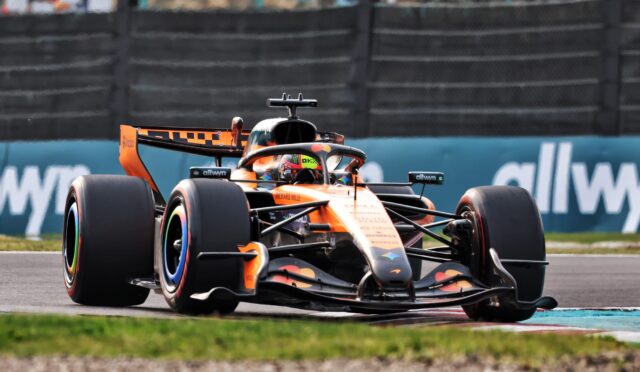 Orange Formula 1 race car speeding through a corner on the track, driver visible in the cockpit wearing a helmet, with barriers in the background.