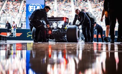 Formula 1 pit crew pushing a race car on a glossy pit lane with a full stands crowd in the background, seen from rear/side view.