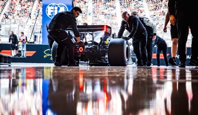 Formula 1 pit crew pushing a race car on a glossy pit lane with a full stands crowd in the background, seen from rear/side view.