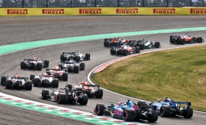 Pack of Formula 1 cars racing through a tight corner, green runoff ahead, with yellow Pirelli barriers and spectators in the stands above.
