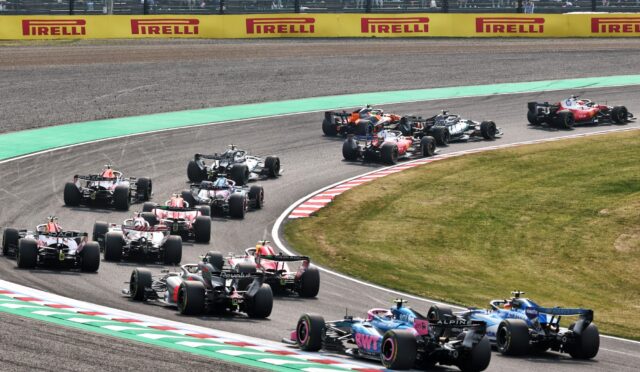 Pack of Formula 1 cars racing through a tight corner, green runoff ahead, with yellow Pirelli barriers and spectators in the stands above.