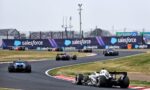 White and black Formula 1 car leads a curve on a race track with Salesforce banners along the barriers and spectators in the stands behind.