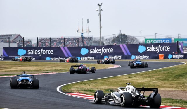 White and black Formula 1 car leads a curve on a race track with Salesforce banners along the barriers and spectators in the stands behind.