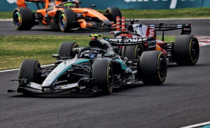 Three Formula 1 cars race along a track, led by a Mercedes in front of a red‑bull style car and an orange McLaren in the background.