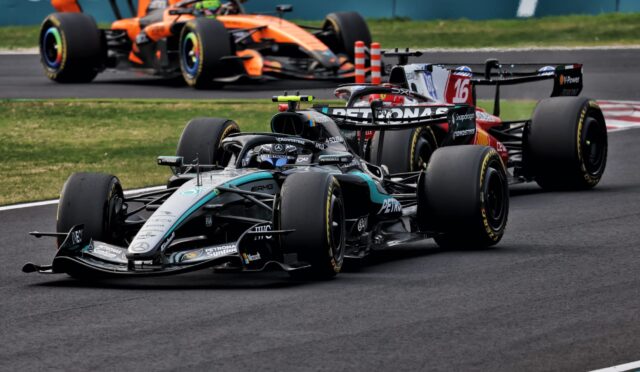Three Formula 1 cars race along a track, led by a Mercedes in front of a red‑bull style car and an orange McLaren in the background.