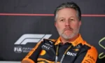 Portrait of a man with gray hair and trimmed beard wearing an orange and black racing team shirt at a Formula 1 backdrop.