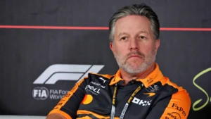 Portrait of a man with gray hair and trimmed beard wearing an orange and black racing team shirt at a Formula 1 backdrop.