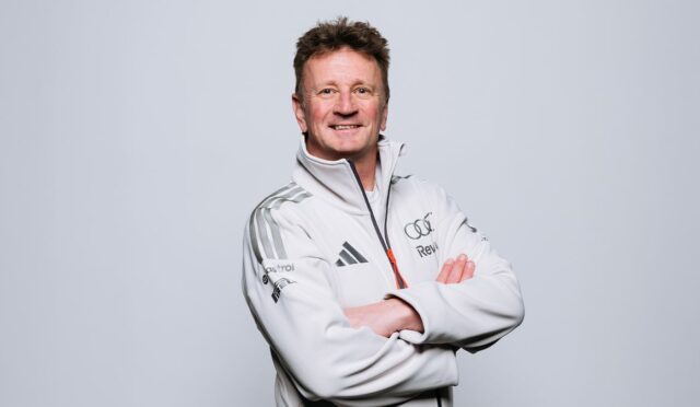 Smiling man in a white athletic jacket with arms crossed, standing in a studio background