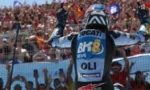 Racing motorcyclist in blue Ducati suit raises both arms in victory on the track, crowd cheering in the stands behind.