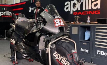 Aprilia Racing motorcycle on a paddock stand in a workshop, front with race number 31, mechanic standing in the background by tool cabinets.