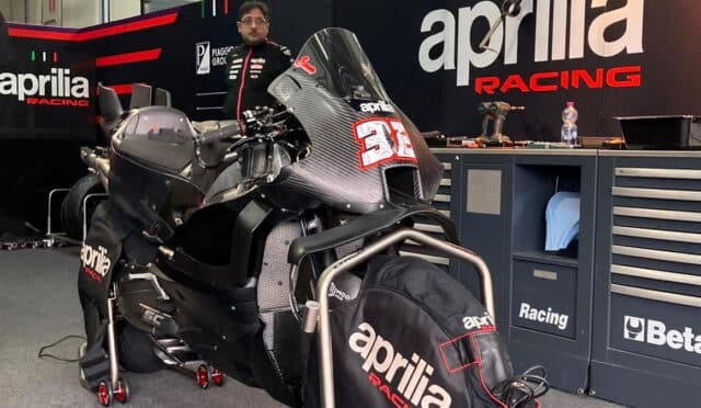 Aprilia Racing motorcycle on a paddock stand in a workshop, front with race number 31, mechanic standing in the background by tool cabinets.