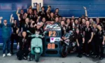 Racing team photo: crowd of riders and crew in black shirts pose with a turquoise scooter and a sport bike in a pit garage.