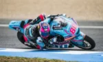 Motorcycle racer wearing turquoise and pink gear leans sharply into a turn on a race track, bike #88 in view.