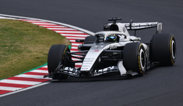 White and black Formula 1 race car corners on a track, driver wearing a blue helmet near red-and-white curbs.