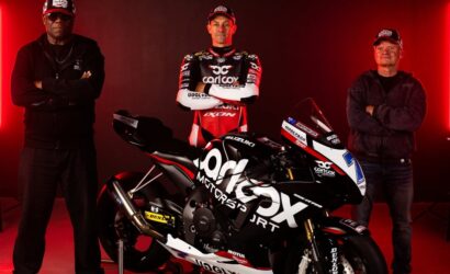 Three men stand on each side of a black Suzuki racing motorcycle in a studio with red neon lights; center man in racing suit poses with arms crossed.
