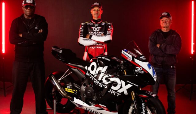 Three men stand on each side of a black Suzuki racing motorcycle in a studio with red neon lights; center man in racing suit poses with arms crossed.