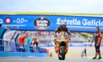 Rider on an orange race bike (number 93) speeds toward the camera on a racing track with a blue Estrella Galicia banner overhead.