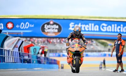 Rider on an orange race bike (number 93) speeds toward the camera on a racing track with a blue Estrella Galicia banner overhead.