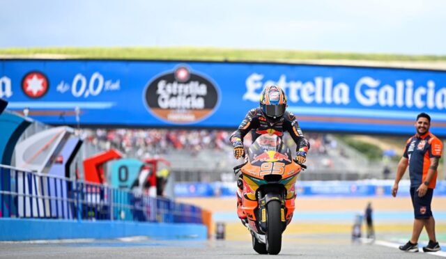 Rider on an orange race bike (number 93) speeds toward the camera on a racing track with a blue Estrella Galicia banner overhead.