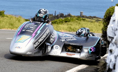 Two riders lean into a sharp turn on a silver sidecar racing motorcycle with colorful sponsor decals, along a coastal road.