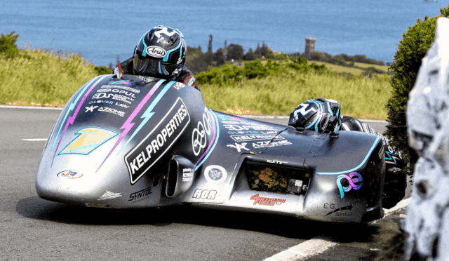 Two riders lean into a sharp turn on a silver sidecar racing motorcycle with colorful sponsor decals, along a coastal road.