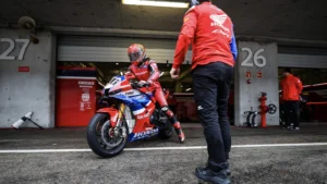 Rider in red racing suit sits on a blue-red motorcycle in the pit lane beside garages 26 and 27, with a crew member nearby.
