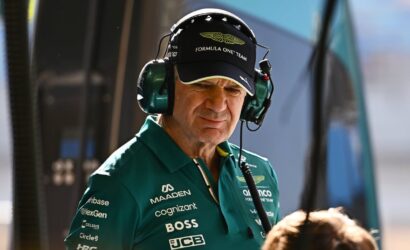 Pit crew member in a green polo and cap with 'Formula One Team' branding, wearing a headset in a pit area, focused on the task
