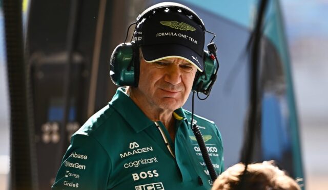 Pit crew member in a green polo and cap with 'Formula One Team' branding, wearing a headset in a pit area, focused on the task
