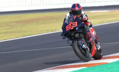 Motorcycle racer in red and black gear leans into a turn on a race track, bike numbered in orange.