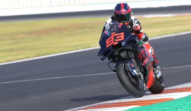 Motorcycle racer in red and black gear leans into a turn on a race track, bike numbered in orange.