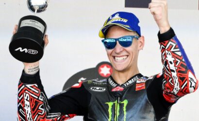 Motocross rider wearing a blue cap and sunglasses celebrates on stage, holding a trophy aloft and smiling broadly.